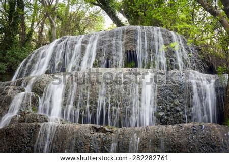 Waterfall among rocks invaded by greenery. Summer sunny, natural park.
