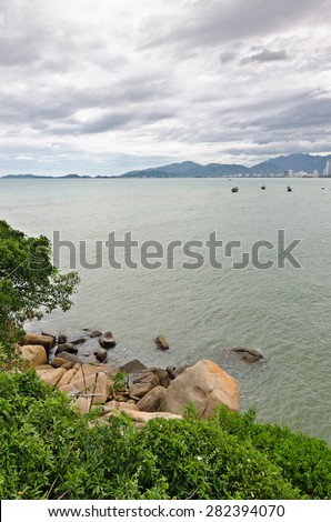 A sea view on a small city from green island with big orange rocks. There are several fishing boats at water.