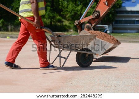 Worker with a cart full of sand. In the background is excavator. Photo was taken on a nice sunny day, at construction site.