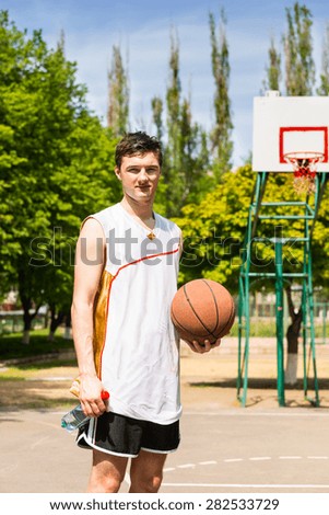 Portrait of Young Athletic Man Standing on Basketball Court Holding Ball on Bright Sunny Day with Backboard and Basket in Background