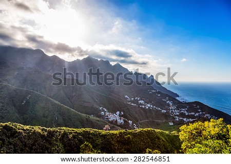 Green mountains or rock with clouds and sun with sunlight and sunbeams on sky over small village or city near the Atlantic ocean in Tenerife Canary island, Spain