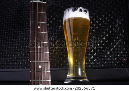 Tall glass full of light beer standing near a grilled music monitor with a guitar fretboard in the foreground Royalty-Free Stock Photo #282566591