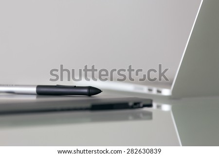 Still life of digital graphic tablet and stylus pen on desk, with laptop computer in background. Reflections on table and copy space on top gray wall
