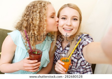 Love my friend. Two attractive young women sitting with glasses of cocktails and doing selfie in cafe, one of them kissing another on cheek.