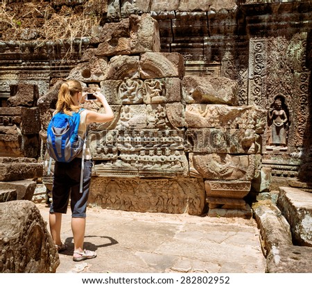 Young female tourist with blue backpack and smartphone taking picture of the bas-reliefs among mysterious ruins of ancient Preah Khan temple in Angkor. Siem Reap, Cambodia.