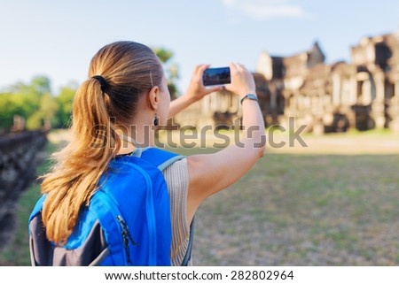 Closeup view of young female tourist with backpack taking picture with smartphone of facade of the ancient mysterious temple complex Angkor Wat on blue sky background. Siem Reap, Cambodia.