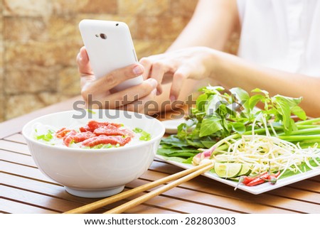 Young woman eating the Pho Bo and using smartphone in street cafe of Vietnam. The Pho Bo is a traditional Vietnamese beef noodle soup with garnish of leaves of cilantro and Asian basil. Popular food.