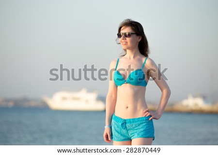 Portrait of young happy woman in sunglasses standing at sea shore with breeze blowing her hair, relaxing in sunlight on sunny beach, white yacht on the background