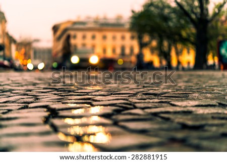 City central square paved with stone after a rain, headlights from cars in the distance. View from the pavement level