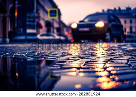 City central square paved with stone after a rain, headlights from car driving on the road. View from the pavement level, image in the orange-blue toning