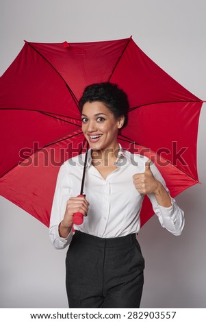 Happy african american business woman standing with red umbrella gesturing thumb up, over gray background