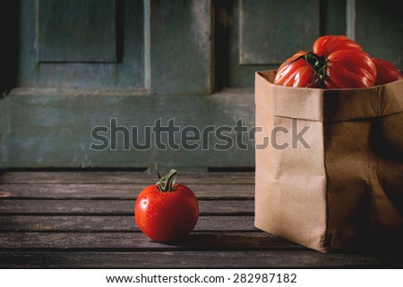 One and heap of big red tomatoes RAF in paper bag over old wooden table. Dark rustic atmosphere