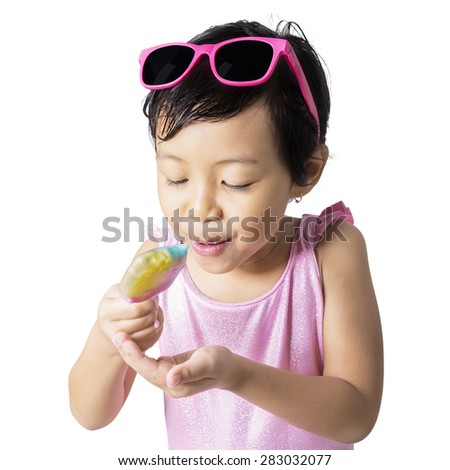 Portrait of little child wearing sunglasses and swimsuit with pink color, standing in the studio while eating a fresh ice cream