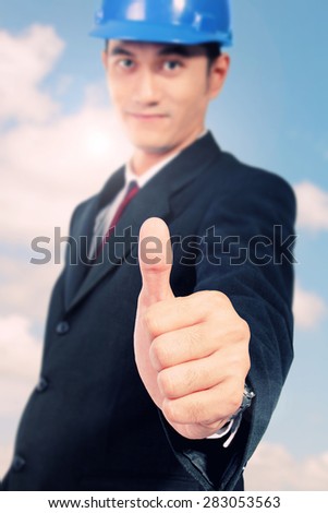 Attractive Asian architect in formal wear giving thumb up, on blue sky background. Conceptual image of trust and safety