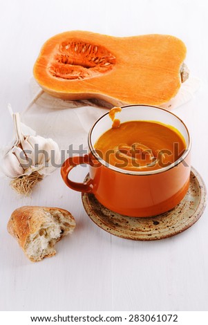 Bowl of creamy pumpkin soup with garlic and bread in a rustic ceramic plate on a white wooden background