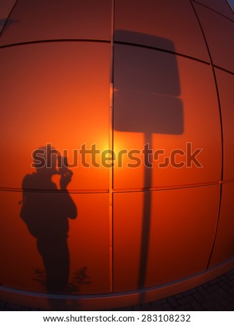 The shadow of a man on a red-orange wall, who photographs a road sign in the evening at sunset with wide angle fisheye lens and distortion view