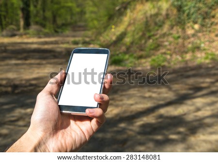 Front view of hand holding black smartphone with white blank screen in forest.