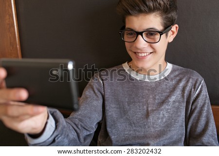 Portrait of smiling teenager man sitting on his bed at home relaxing, holding a smartphone networking on-line, taking selfies portraits of himself wearing glasses, interior. Lifestyle technology. 