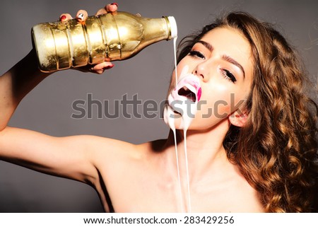 Impassioned attractive young woman with curly hair holding gold bottle pours white yogurt on her face with open mouth looking forward standing on grey background, horizontal picture