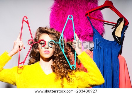 Modern young girl with curly hair in yellow sweater holding hangers standing amid colorful clothes pink red blue colors on grey wall background, horizontal picture