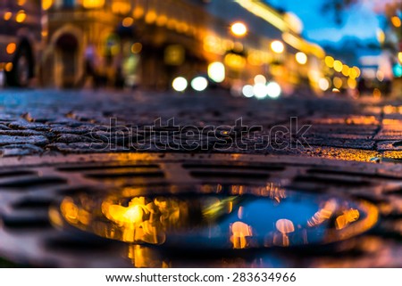 The bright lights of the evening city after rain, headlights from cars in the distance. View from the hatch on the pavement level