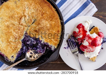 Homemade fresh blueberry cobbler baked in cast iron skillet with large spoon and white plate with single sliced topped with vanilla ice cream and fresh strawberry compote