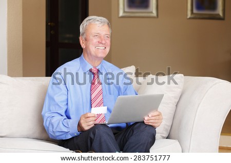 Portrait of retired senior man sitting at sofa while holding credit card in his hands and shopping online with laptop. 