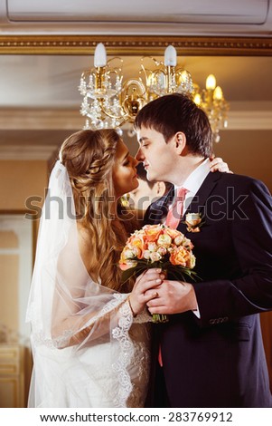 Beautiful loving wedding couple is holding flowers bouquet and kissing at luxury restaurant interior background.