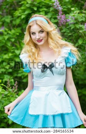 Portrait of a beautiful young blonde woman with long hair dressed as Alice in Wonderland. Girl on the nature near the lilac bushes. Soft focus 