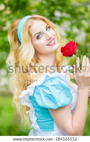 Portrait of a beautiful young blonde woman with long hair dressed as Alice in Wonderland.The girl is holding a red rose. Soft focus 