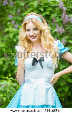 Portrait of a beautiful young blonde woman with long hair dressed as Alice in Wonderland. Girl on the nature near the lilac bushes. Soft focus 