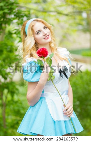 Portrait of a beautiful young blonde woman with long hair dressed as Alice in Wonderland.The girl is holding a red rose. Soft focus 