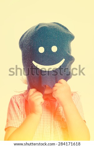 Little girl with a face covered by a blue hat with a happy smiley, isolated