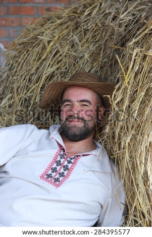 a man with a black beard in a white embroidered shirt lies near the sheaf of straw