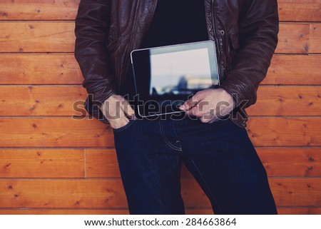 Cropped image with stylish young man holding digital tablet with a blank screen, hipster student with touch pad standing on wooden background, man's hands holding black tablet with copy space area