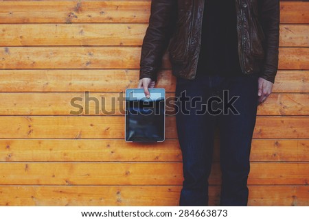 Cropped image with stylish young man holding blank digital tablet while standing on wooden background with big copy space area for text message or content, male hipster hand holding touch pad, filter