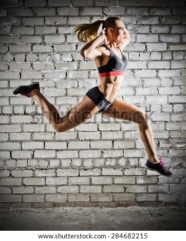 Muscular jumping woman on grey brick wall background (dark version)