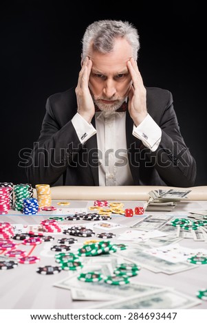 Man in years sitting at poker table and considering poker strategy with his hands on temples. The chips and money are on table