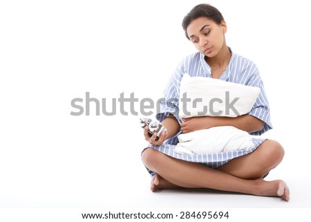 Feeling upset. Portrait of youthful woman sitting white pillow and looking at alarm clock on isolated background