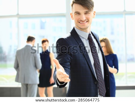 Smiling businessman stretching his hand out for a handshake in foreground and his coworkers discussing business