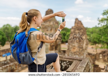 Closeup view of young female tourist with blue backpack taking picture with smartphone from the top of ancient Pre Rup temple Khmer in Angkor. Siem Reap, Cambodia.