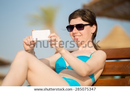 Young happy smiling woman in sunglasses taking self portrait with smartphone while relaxing and tanning on wooden sun lounger on sunny southern beach with straw umbrellas