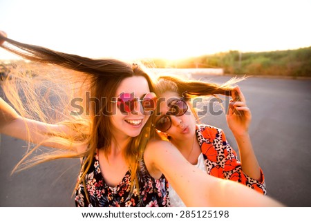 Two attractive young girl friends standing together and posing on camera.Brunette having fun and showing sign with her hand.photography at sunset.two women taking selfie with mobile phone with fisheye