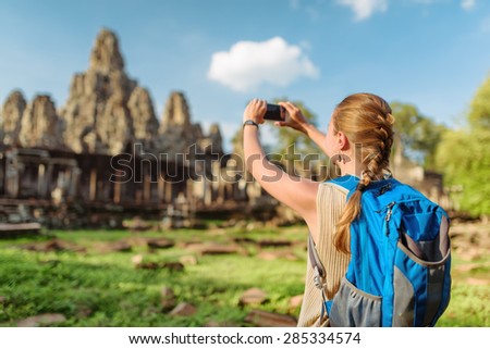 Young female tourist with backpack and smartphone taking picture of ancient Bayon temple in Angkor Thom on blue sky background in evening sun. Siem Reap, Cambodia.