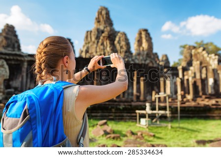 Closeup view of young female tourist with blue backpack and smartphone taking picture among mysterious ruins of ancient Angkor Thom in Siem Reap, Cambodia. Bayon temple on blue sky background.