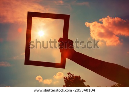 Hand holding a wooden frame on sunrise sky background. Care, safety, memory or painting concept.