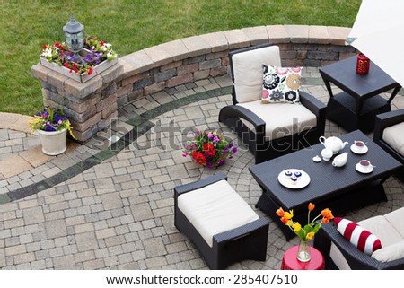Brick paved patio with comfortable patio furniture with modern armchairs and a stool around a table set with tea and cookies alongside a low curving wall overlooking a green lawn, high angle view