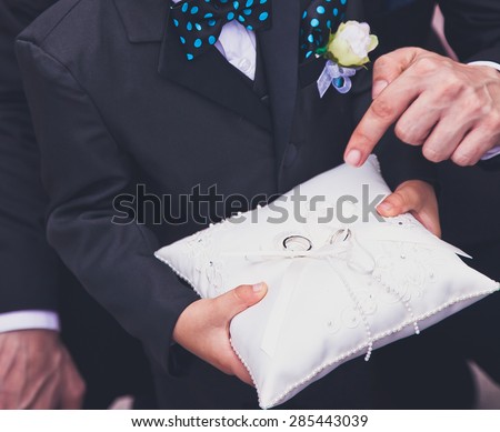Young boy carrying wedding rings on a pillow during wedding ceremony and his father helps him and points at rings. Boy in a suit and with a rose and bow tie Royalty-Free Stock Photo #285443039