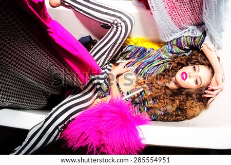 Spectacular emotional young woman with curly hair lying in white bathtub amid colorful clothes pink orange red blue colors on grey wall background, horizontal picture