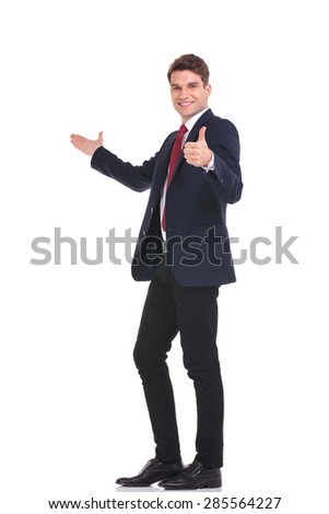 Full body picture of a young handsome business man presenting while showing the thumbs up gesture.